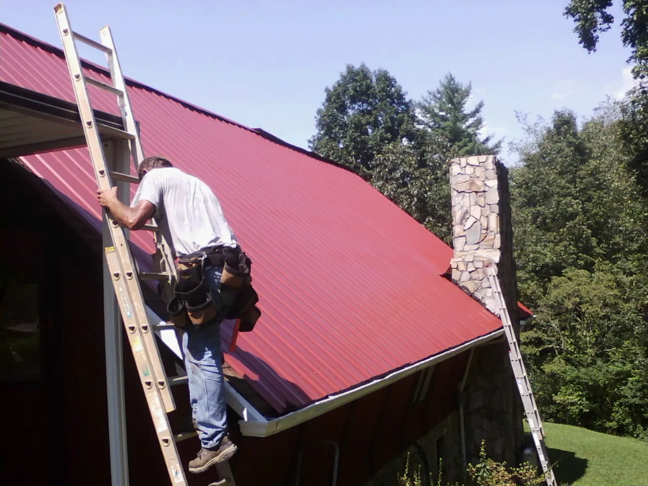 Roofer on a ladder beside a red metal roof and stone chimney — Soffit & Fascia Repair by Roofing Brothers in Saint Meinrad